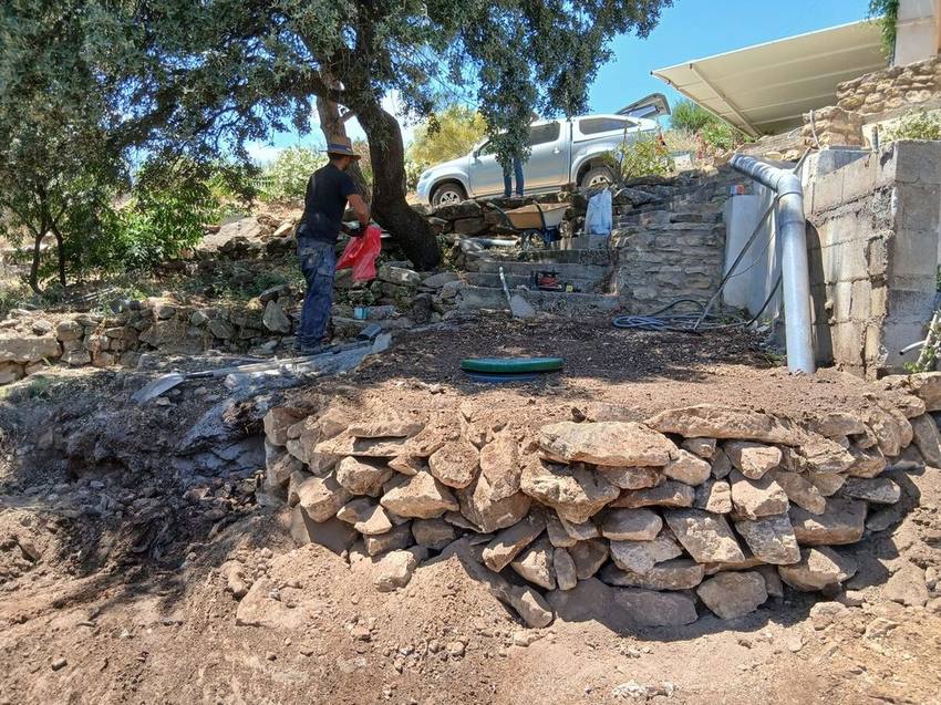 Operario trabajando en un terreno en obra junto a un muro de piedra, con una tapa circular verde en el suelo y una tubería gris